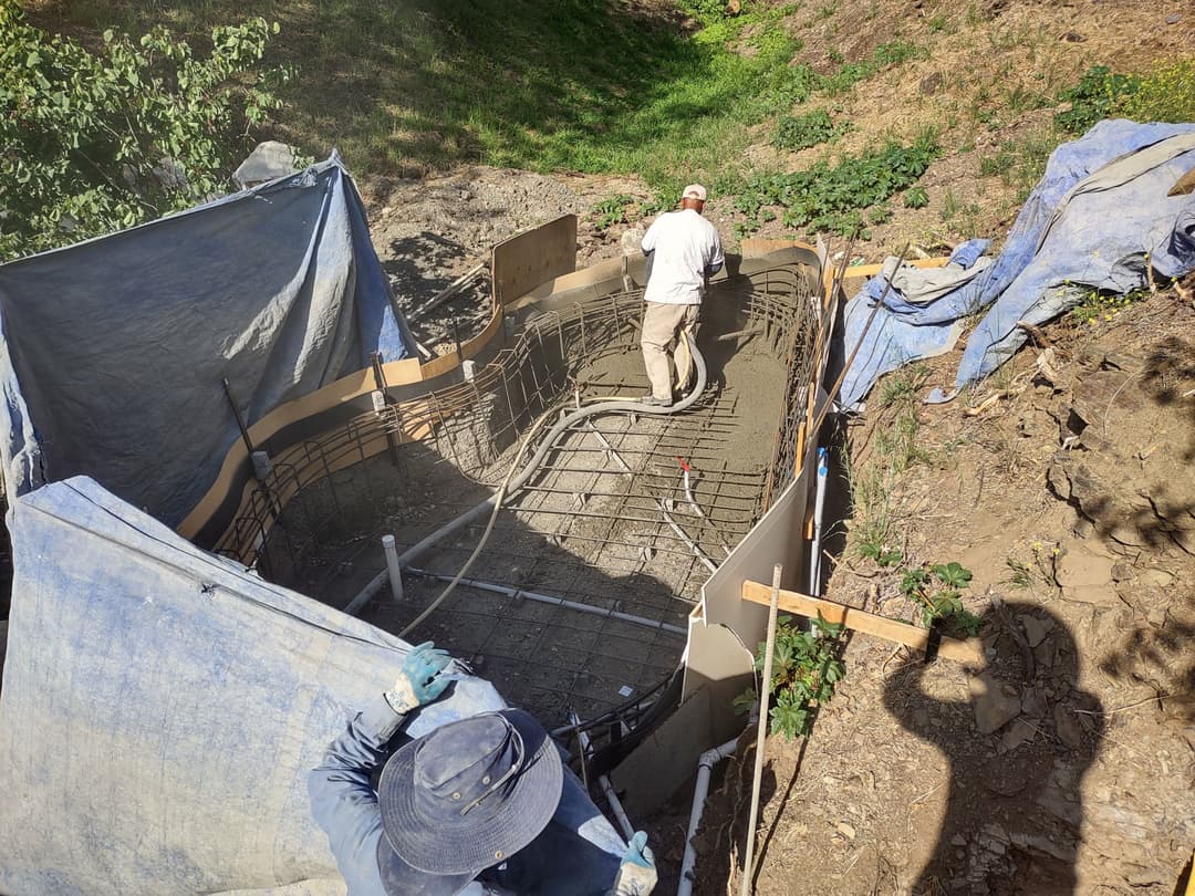 Construction of a concrete pool with workers in a dirt setting, surrounded by protective tarps.
