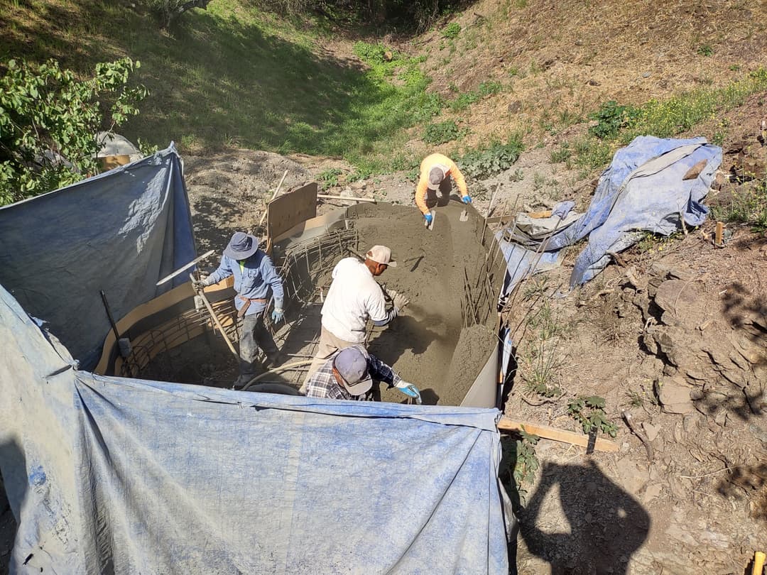 Workers pouring concrete into a foundation form on a construction site surrounded by tarps.