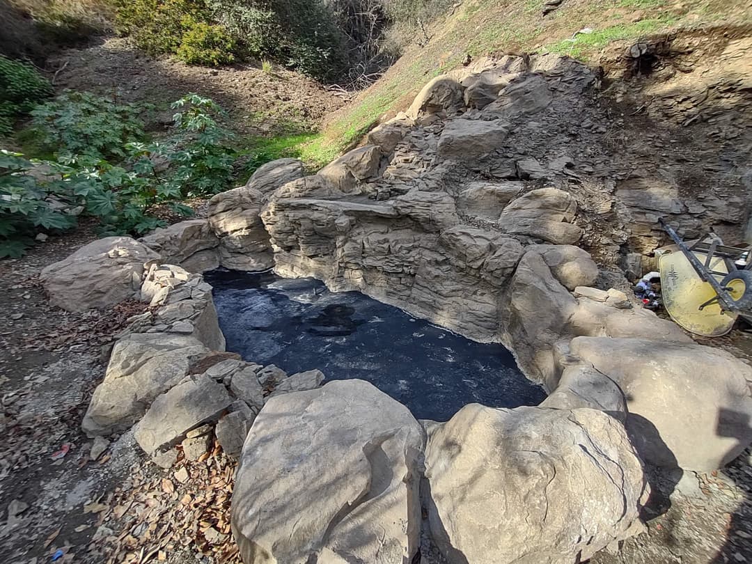Natural stone hot spring pool surrounded by greenery and rocky terrain.
