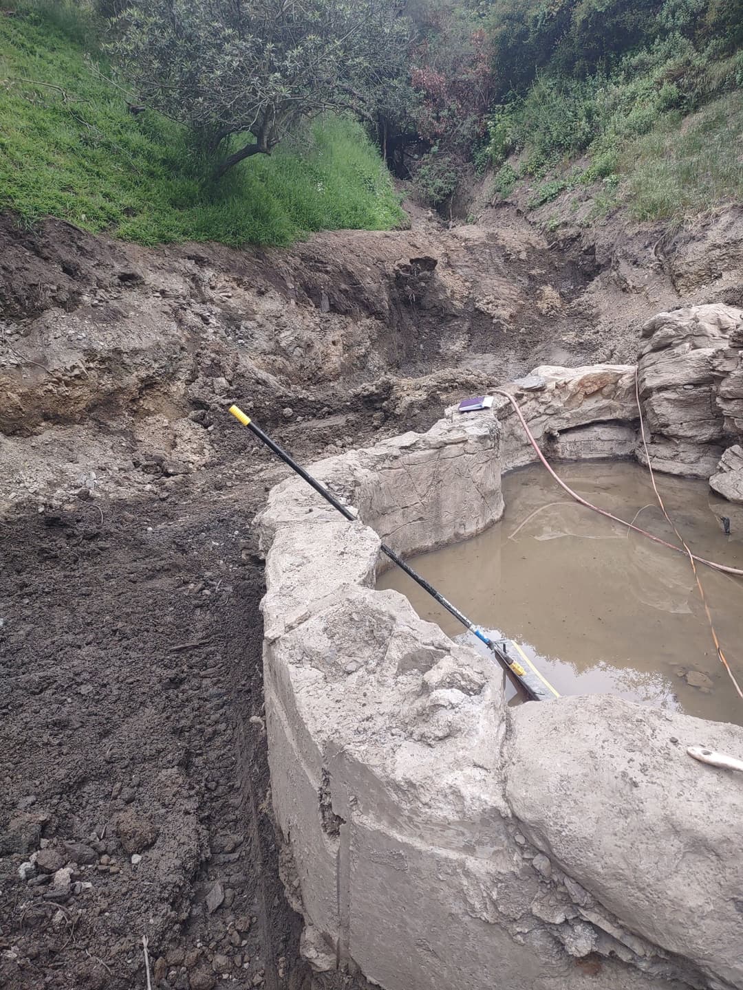 Excavation site with a circular concrete foundation and water-filled pit surrounded by dirt.