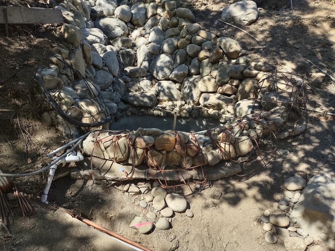 Stone water catchment basin under construction with metal reinforcement and surrounding rocks.