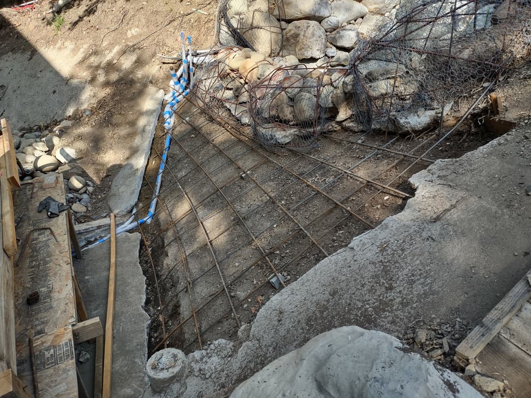 Concrete foundation with rebar grid, rocky surroundings, and construction materials nearby.