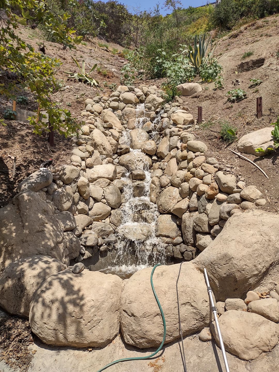 Rocky waterfall feature in a garden, surrounded by greenery and natural landscaping.