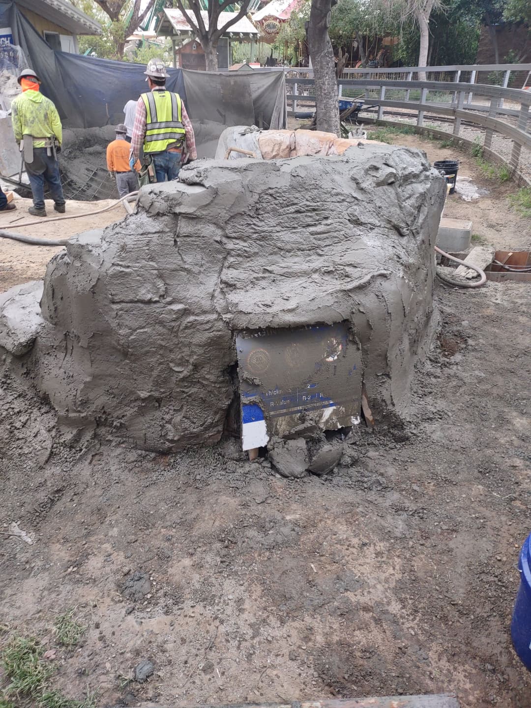 Construction site with workers applying wet concrete to a curved structure. Tools and materials visible.