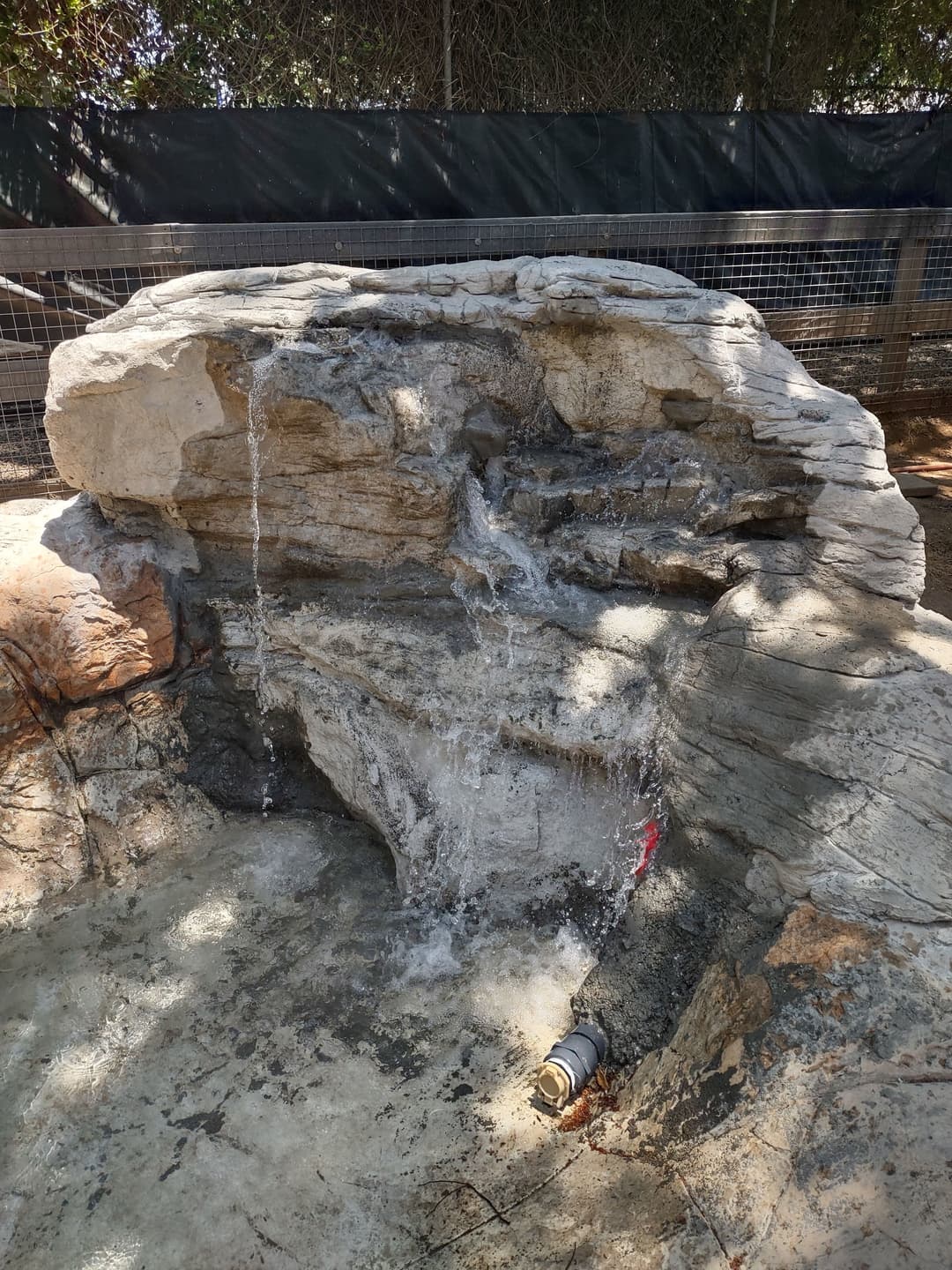 Artificial rock waterfall with water flowing, surrounded by a fenced area in a park setting.