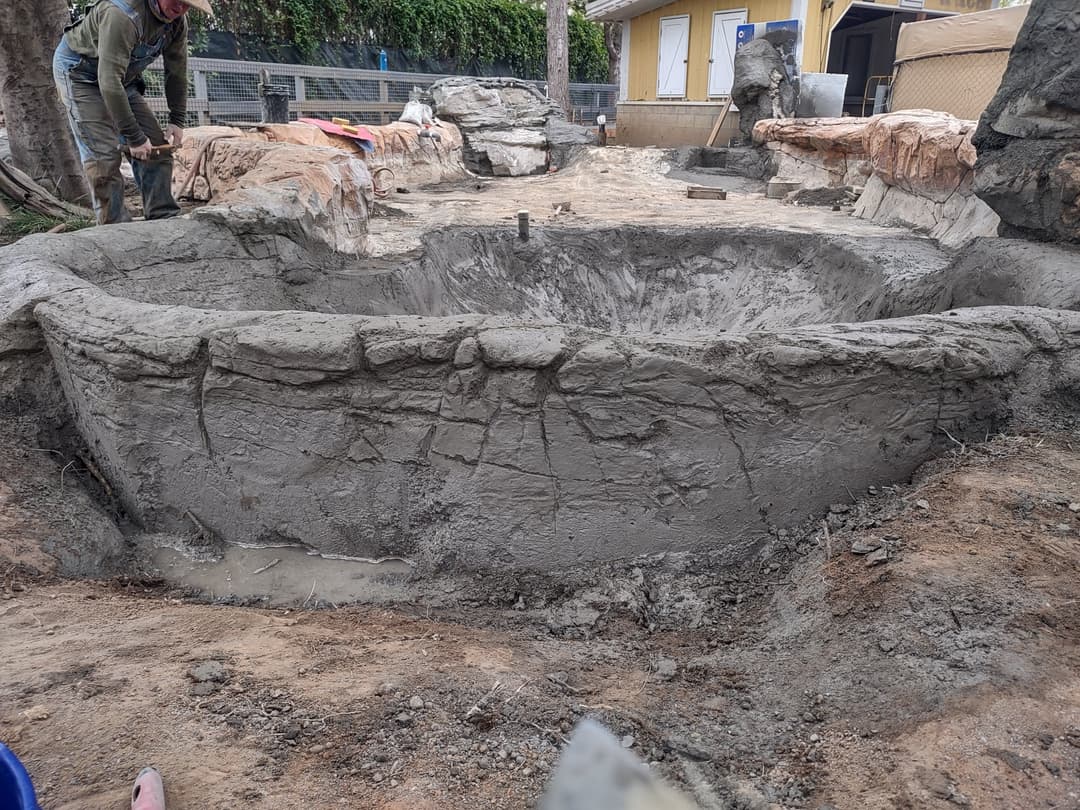 Excavated pond under construction with rough stone edges and tools nearby.