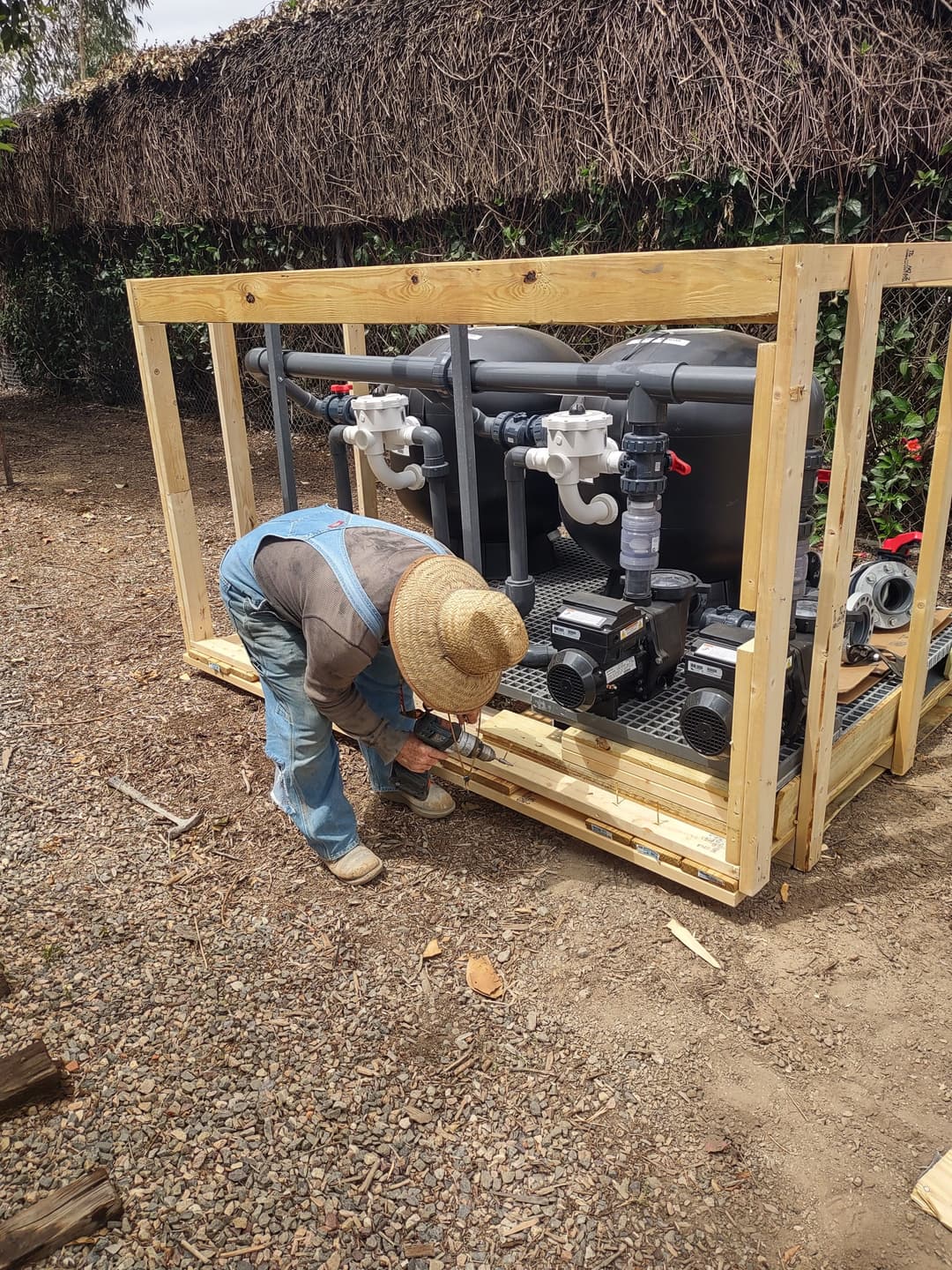 Worker assembling pool equipment in a wooden frame outdoors with tools and machinery visible.