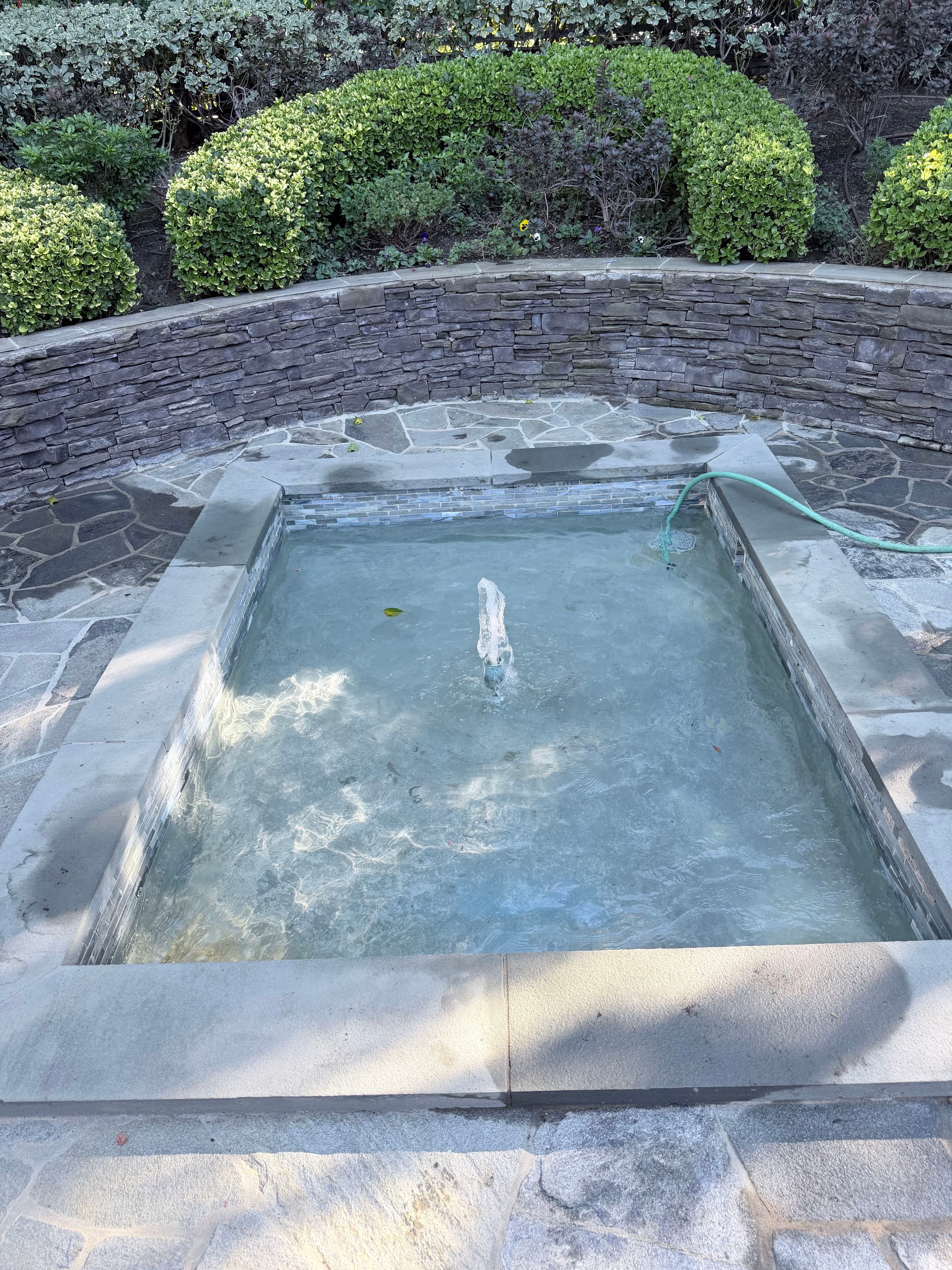 Garden fountain surrounded by stone pathway and lush greenery, gently flowing water.