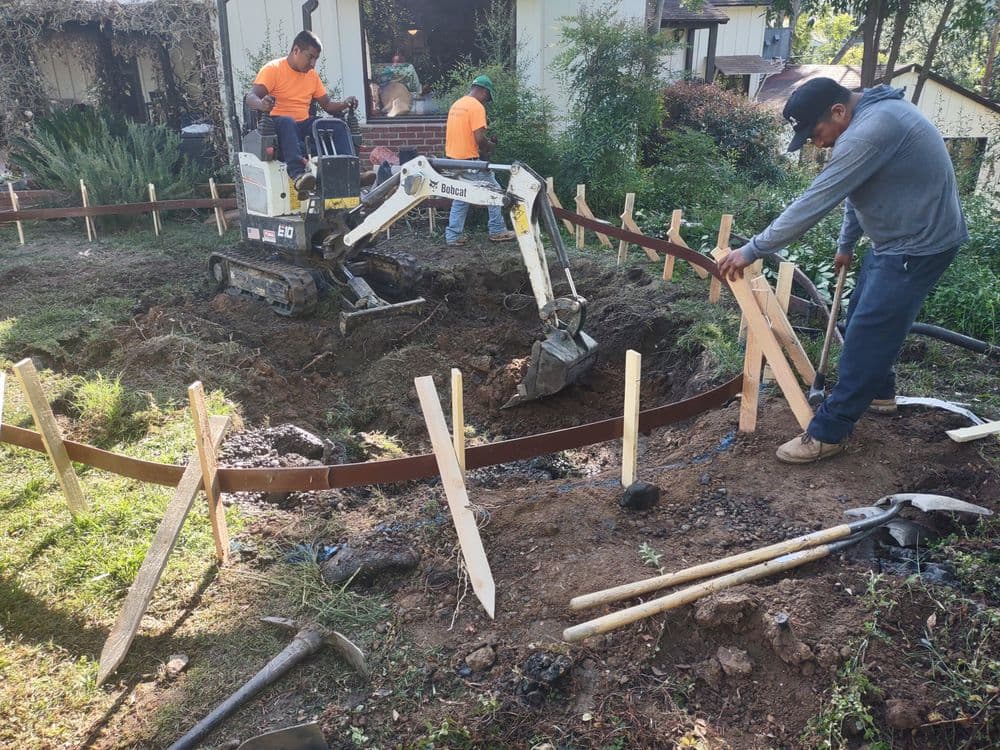 Workers using heavy machinery to create a foundation in a garden area with wooden stakes.